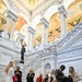 A group of approximately ten visitors are gathered in the ornate Great Hall of the Library of Congress Thomas Jefferson Building in Washington, D.C. The space features elaborate marble staircases, Corinthian columns, richly painted vaulted ceilings in gold, red, and green, and a bronze statue of a torch-bearing figure. The visitors appear to be on a guided tour, with some looking upward at the architecture and one person reviewing materials on a tablet.