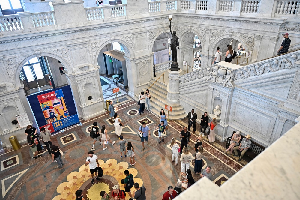 An aerial view of a grand neoclassical building interior, likely the Library of Congress Great Hall in Washington D.C., featuring ornate white marble architecture with arched colonnades and decorative inlaid flooring. The space is populated with numerous visitors milling about the main floor and upper balcony level. A prominent bronze statue on a pedestal is visible at the center-rear of the hall, and a 'National Book Festival' banner is displayed on the left wall.
