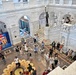 An aerial view of a grand neoclassical building interior, likely the Library of Congress Great Hall in Washington D.C., featuring ornate white marble architecture with arched colonnades and decorative inlaid flooring. The space is populated with numerous visitors milling about the main floor and upper balcony level. A prominent bronze statue on a pedestal is visible at the center-rear of the hall, and a 'National Book Festival' banner is displayed on the left wall.