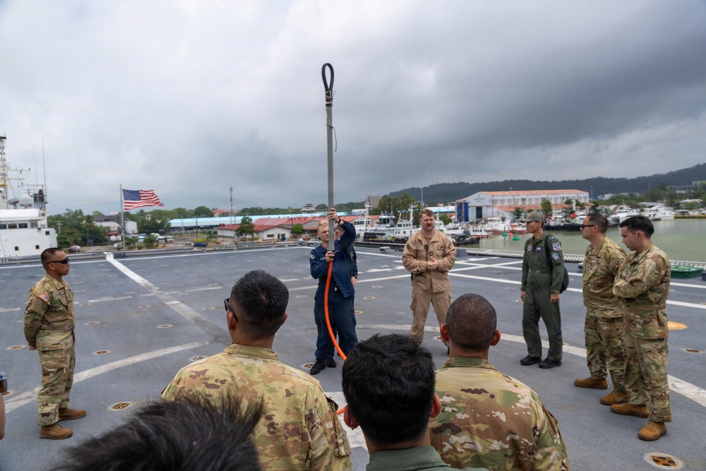 Military personnel from multiple services are gathered on the deck of a naval vessel for what appears to be a training or demonstration session. A person in a blue flight suit is holding up a long pole or tool with an orange rope/cord attached, demonstrating its use to the assembled group. The setting appears to be a port or harbor, with an American flag visible and waterfront facilities in the background.