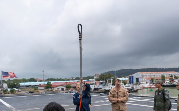 Military personnel from multiple services are gathered on the deck of a naval vessel for what appears to be a training or demonstration session. A person in a blue flight suit is holding up a long pole or tool with an orange rope/cord attached, demonstrating its use to the assembled group. The setting appears to be a port or harbor, with an American flag visible and waterfront facilities in the background.