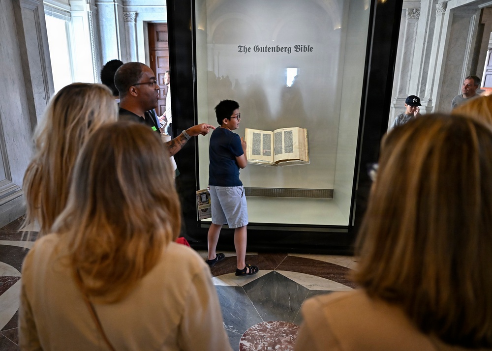 Visitors are gathered around a glass display case containing the Gutenberg Bible at what appears to be a museum or library. A young boy wearing glasses and a blue shirt stands closest to the display case, pointing at the open book. The setting features ornate marble architecture consistent with a major institutional building.