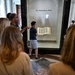 Visitors are gathered around a glass display case containing the Gutenberg Bible at what appears to be a museum or library. A young boy wearing glasses and a blue shirt stands closest to the display case, pointing at the open book. The setting features ornate marble architecture consistent with a major institutional building.