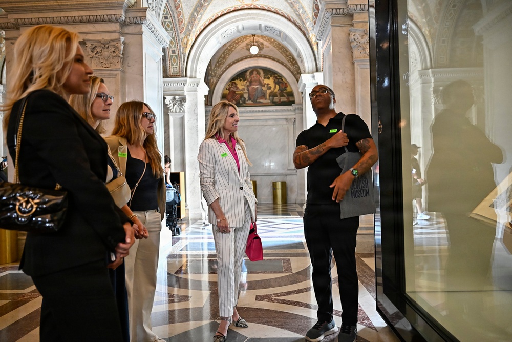 A group of visitors are gathered in an ornate neoclassical building, likely the Library of Congress in Washington D.C., based on the distinctive marble columns, arched ceilings, and decorative murals. The group appears to be on a guided tour or organized visit, with several individuals wearing green visitor badges. They are gathered near a glass display case or partition, with one person appearing to explain or point something out.