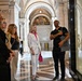 A group of visitors are gathered in an ornate neoclassical building, likely the Library of Congress in Washington D.C., based on the distinctive marble columns, arched ceilings, and decorative murals. The group appears to be on a guided tour or organized visit, with several individuals wearing green visitor badges. They are gathered near a glass display case or partition, with one person appearing to explain or point something out.