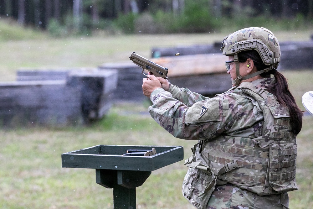 A female soldier in full military combat gear is firing a semi-automatic pistol at an outdoor shooting range. A spent brass casing is visibly ejecting from the firearm, indicating the weapon has just been discharged. The setting appears to be a military training range with target berms visible in the background.