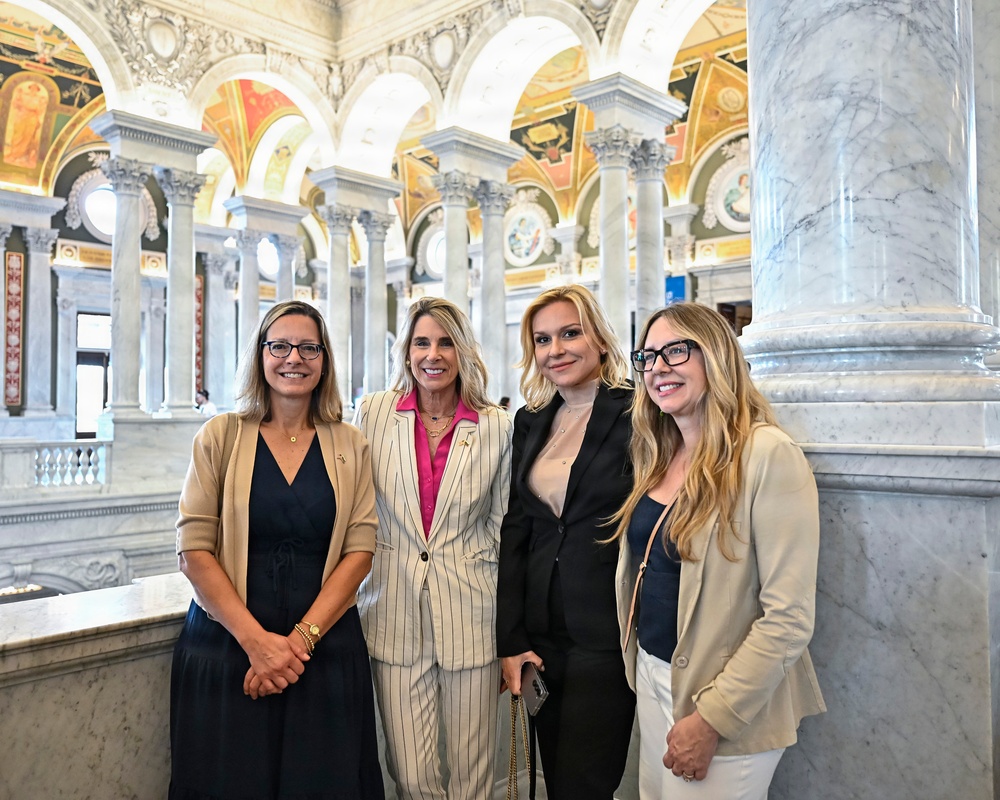 Four women are posing together for a group photo inside an ornate neoclassical building, identifiable as the Library of Congress Great Hall in Washington, D.C. The setting features elaborate marble columns, arched ceilings with colorful murals and gilded decorations. The women are dressed in business-casual to formal attire and appear to be smiling for the camera.