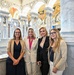 Four women are posing together for a group photo inside an ornate neoclassical building, identifiable as the Library of Congress Great Hall in Washington, D.C. The setting features elaborate marble columns, arched ceilings with colorful murals and gilded decorations. The women are dressed in business-casual to formal attire and appear to be smiling for the camera.