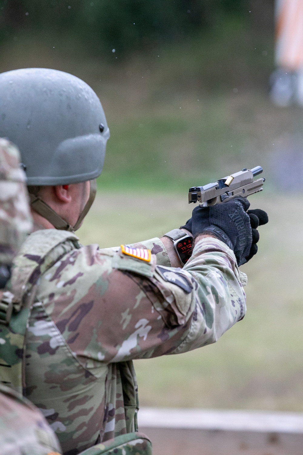 A soldier in MultiCam camouflage uniform and ballistic helmet is firing a semi-automatic pistol at a shooting range. The slide of the pistol appears to be in recoil, indicating the weapon has just been discharged. The soldier is wearing tactical gloves, a smartwatch, and has unit patches visible on the left shoulder.