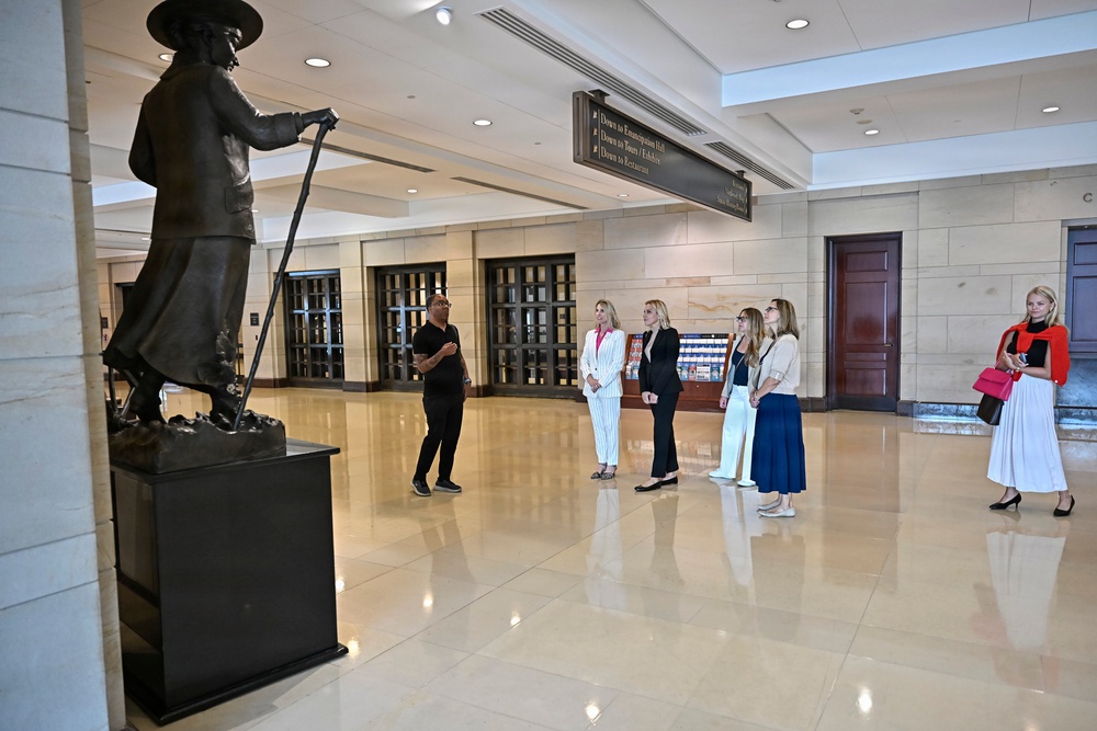 A group of people are gathered in what appears to be a formal government or museum building lobby, listening to a guide or docent who is presenting near a large bronze statue. The statue depicts a historical figure wearing a wide-brimmed hat and holding a long implement, mounted on a dark pedestal. The setting features marble floors, stone walls, and directional signage overhead.