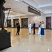 A group of people are gathered in what appears to be a formal government or museum building lobby, listening to a guide or docent who is presenting near a large bronze statue. The statue depicts a historical figure wearing a wide-brimmed hat and holding a long implement, mounted on a dark pedestal. The setting features marble floors, stone walls, and directional signage overhead.