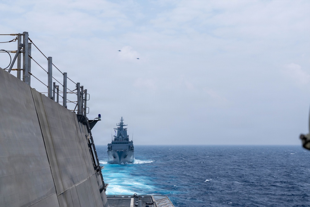 A naval formation at sea showing two warships sailing in close proximity, photographed from the deck of one vessel looking toward a second warship ahead. Two aircraft are visible flying in formation in the overcast sky above. The scene appears to depict a multinational naval exercise or coordinated patrol operation.
