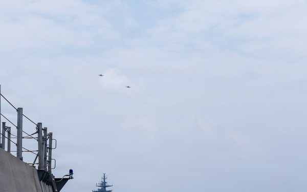 A naval formation at sea showing two warships sailing in close proximity, photographed from the deck of one vessel looking toward a second warship ahead. Two aircraft are visible flying in formation in the overcast sky above. The scene appears to depict a multinational naval exercise or coordinated patrol operation.