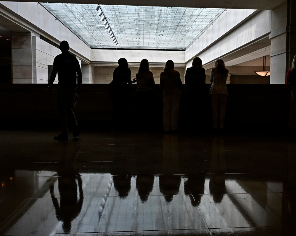 The image shows silhouetted figures in what appears to be a large institutional or public building interior, backlit by a prominent glass skylight ceiling. Several people are standing at what appears to be a counter or railing, while one individual walks separately to the left. The highly polished floor creates strong mirror-like reflections of the figures and architectural elements.