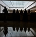 The image shows silhouetted figures in what appears to be a large institutional or public building interior, backlit by a prominent glass skylight ceiling. Several people are standing at what appears to be a counter or railing, while one individual walks separately to the left. The highly polished floor creates strong mirror-like reflections of the figures and architectural elements.