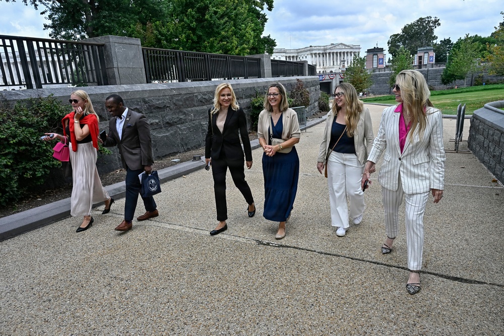 A group of six professionally dressed individuals are walking together along a paved pathway near what appears to be the U.S. Capitol building in Washington, D.C. The group consists of five women and one man, all dressed in business or business-casual attire. The setting suggests an official or formal occasion in a government district.