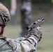 A military service member in full combat gear is firing a semi-automatic pistol at a shooting range. The slide of the pistol is visibly cycling, with a brass cartridge casing being ejected mid-shot. The shooter is wearing MultiCam camouflage uniform and a tactical helmet, suggesting a training exercise.