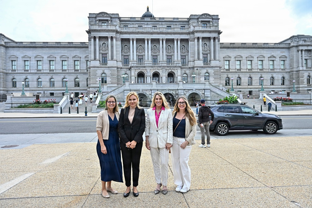 Four women pose for a group photo in front of the Library of Congress Thomas Jefferson Building in Washington, D.C. They are dressed in professional business attire and appear to be visiting or working in the Capitol Hill area. The neoclassical architecture of the landmark building is clearly visible in the background.