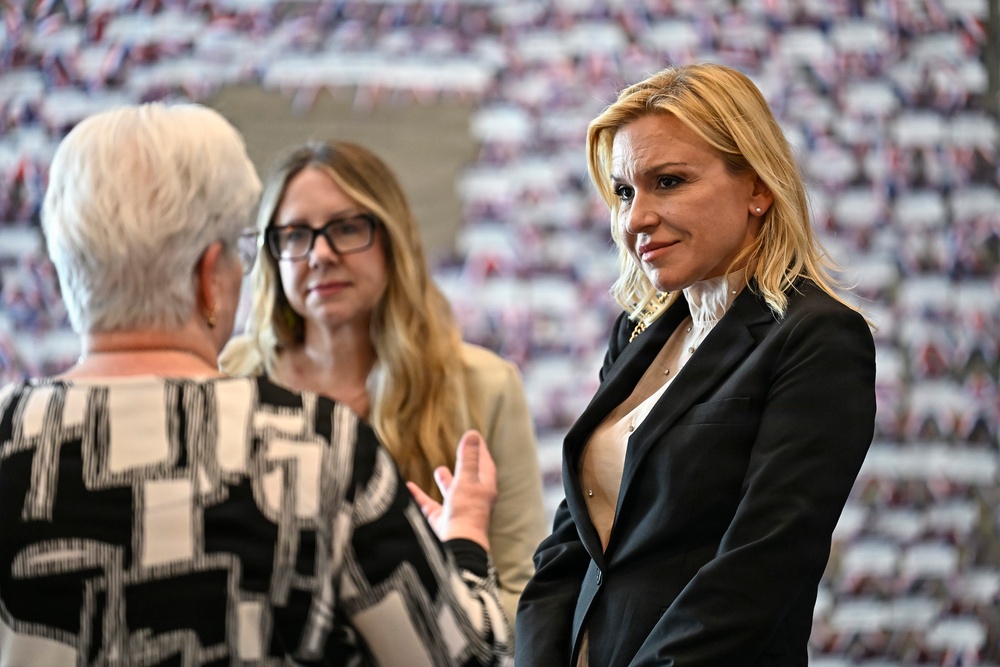 Three women are engaged in conversation at what appears to be a formal or political event. The woman on the right, wearing a black blazer, is attentively listening while the others speak. The background features a large display of repeated images, possibly flags or crowd photos.