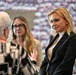 Three women are engaged in conversation at what appears to be a formal or political event. The woman on the right, wearing a black blazer, is attentively listening while the others speak. The background features a large display of repeated images, possibly flags or crowd photos.