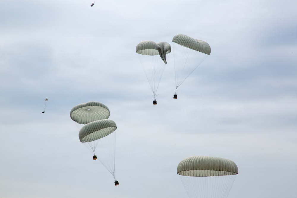 The image shows multiple military-style cargo parachutes descending through an overcast sky. The parachutes are olive/sage green in color and appear to be carrying supply crates or equipment bundles suspended beneath them. This appears to be a military airdrop or supply delivery operation with at least six parachutes visible at various altitudes.