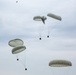 The image shows multiple military-style cargo parachutes descending through an overcast sky. The parachutes are olive/sage green in color and appear to be carrying supply crates or equipment bundles suspended beneath them. This appears to be a military airdrop or supply delivery operation with at least six parachutes visible at various altitudes.