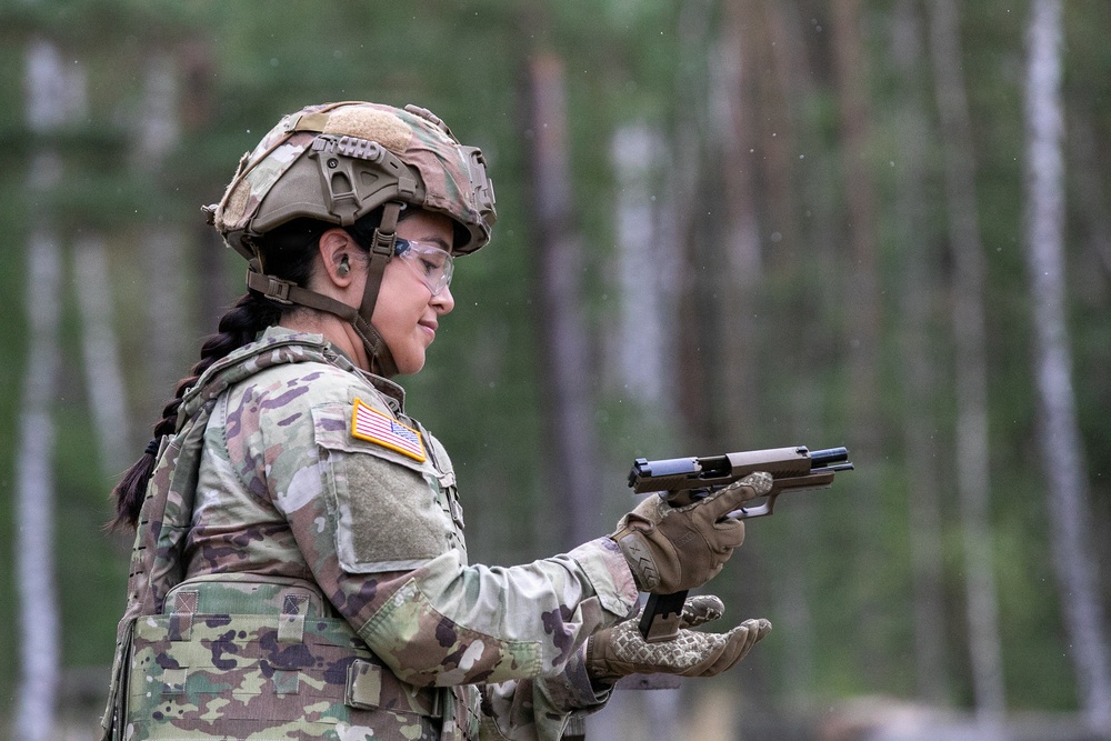 A female U.S. Army soldier in full combat gear is handling a semi-automatic pistol, appearing to inspect or clear the weapon. She is wearing Operational Camouflage Pattern (OCP) uniform with a U.S. flag patch, tactical helmet, protective eyewear, tactical gloves, and a body armor vest. The setting appears to be an outdoor training range surrounded by trees.