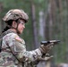A female U.S. Army soldier in full combat gear is handling a semi-automatic pistol, appearing to inspect or clear the weapon. She is wearing Operational Camouflage Pattern (OCP) uniform with a U.S. flag patch, tactical helmet, protective eyewear, tactical gloves, and a body armor vest. The setting appears to be an outdoor training range surrounded by trees.