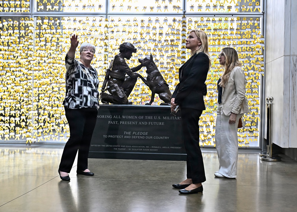 Three women stand near a bronze military sculpture in what appears to be a memorial or museum setting. The sculpture depicts a female soldier with a military working dog, mounted on a plaque honoring women of the U.S. Military. The background wall is covered with numerous yellow ribbons, each appearing to contain individual dedications or names.