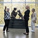Three women stand near a bronze military sculpture in what appears to be a memorial or museum setting. The sculpture depicts a female soldier with a military working dog, mounted on a plaque honoring women of the U.S. Military. The background wall is covered with numerous yellow ribbons, each appearing to contain individual dedications or names.
