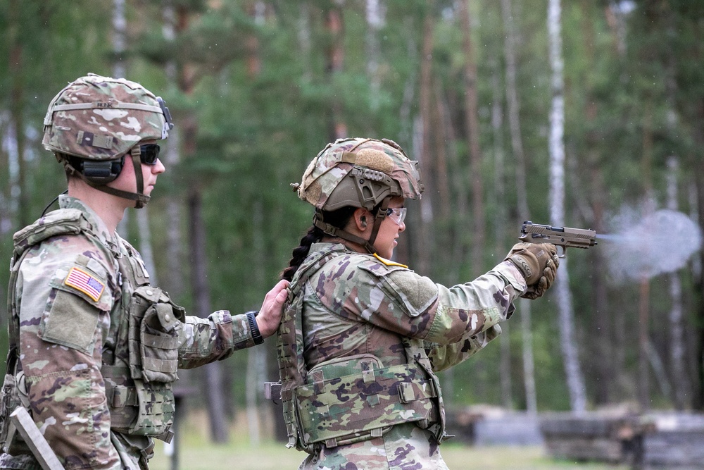 Two U.S. Army soldiers are at an outdoor firing range, with one soldier actively firing a pistol while the other observes and provides oversight. The shooter, wearing a tan tactical helmet and protective eyewear, has just discharged the weapon as evidenced by visible muzzle smoke. The setting appears to be a wooded training area, consistent with a military marksmanship qualification or training exercise.