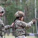 Two U.S. Army soldiers are at an outdoor firing range, with one soldier actively firing a pistol while the other observes and provides oversight. The shooter, wearing a tan tactical helmet and protective eyewear, has just discharged the weapon as evidenced by visible muzzle smoke. The setting appears to be a wooded training area, consistent with a military marksmanship qualification or training exercise.