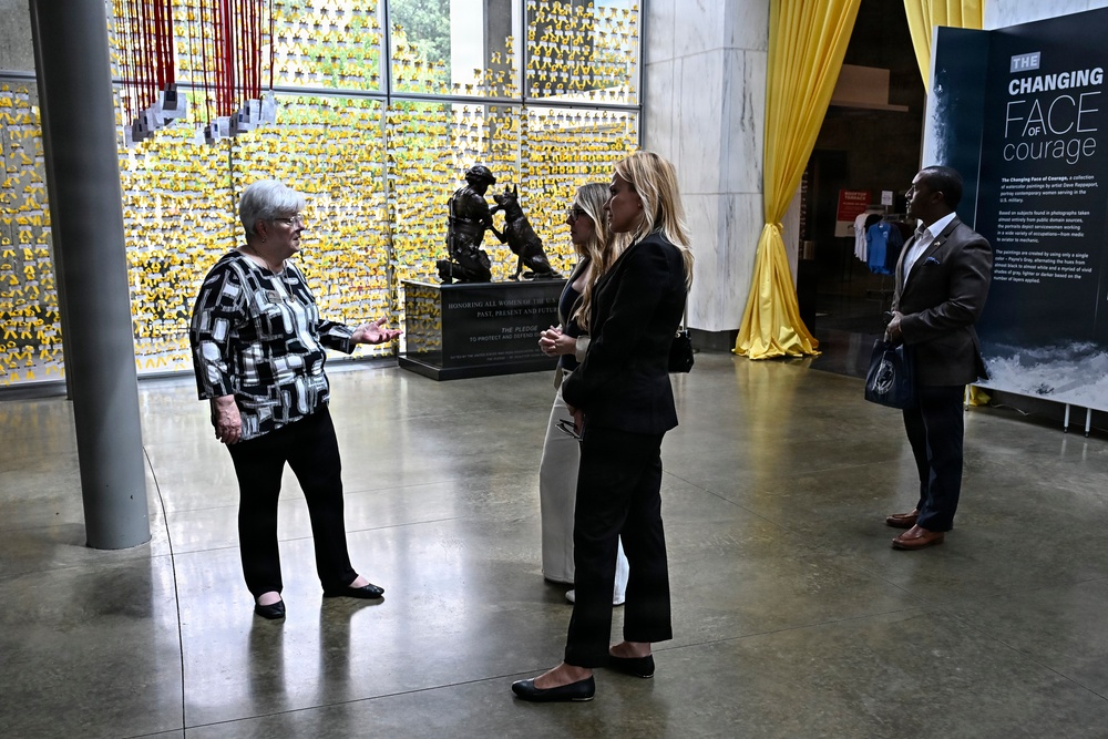 Four individuals are gathered in what appears to be a museum or memorial space, engaged in conversation. The setting features a prominent wall covered in yellow ribbons and a bronze sculpture with a dedication plaque honoring women of the U.S. military. A banner on the right promotes an exhibition titled 'The Changing Face of Courage.'
