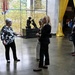 Four individuals are gathered in what appears to be a museum or memorial space, engaged in conversation. The setting features a prominent wall covered in yellow ribbons and a bronze sculpture with a dedication plaque honoring women of the U.S. military. A banner on the right promotes an exhibition titled 'The Changing Face of Courage.'