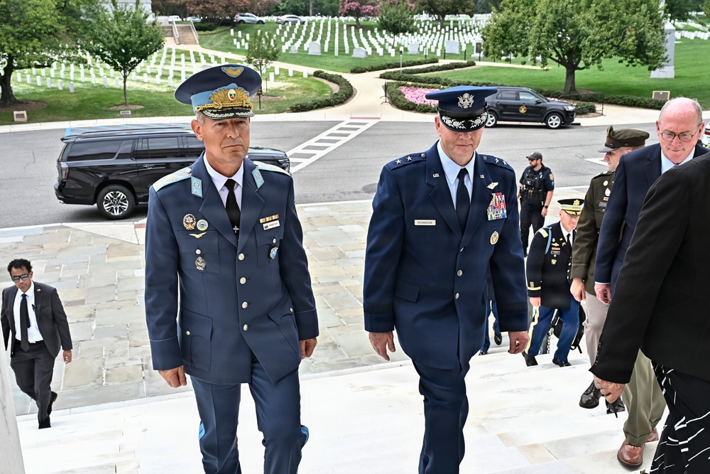 Two senior military officers in dress uniforms ascend stone steps at what appears to be Arlington National Cemetery, accompanied by a group of officials and security personnel. The officer on the left wears a light blue foreign military uniform with numerous decorations and a distinctive peaked cap, while the officer on the right wears a US Air Force dress uniform with a major general rank insignia. White grave markers are visible in the background, confirming the cemetery setting.