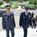 Two senior military officers in dress uniforms ascend stone steps at what appears to be Arlington National Cemetery, accompanied by a group of officials and security personnel. The officer on the left wears a light blue foreign military uniform with numerous decorations and a distinctive peaked cap, while the officer on the right wears a US Air Force dress uniform with a major general rank insignia. White grave markers are visible in the background, confirming the cemetery setting.