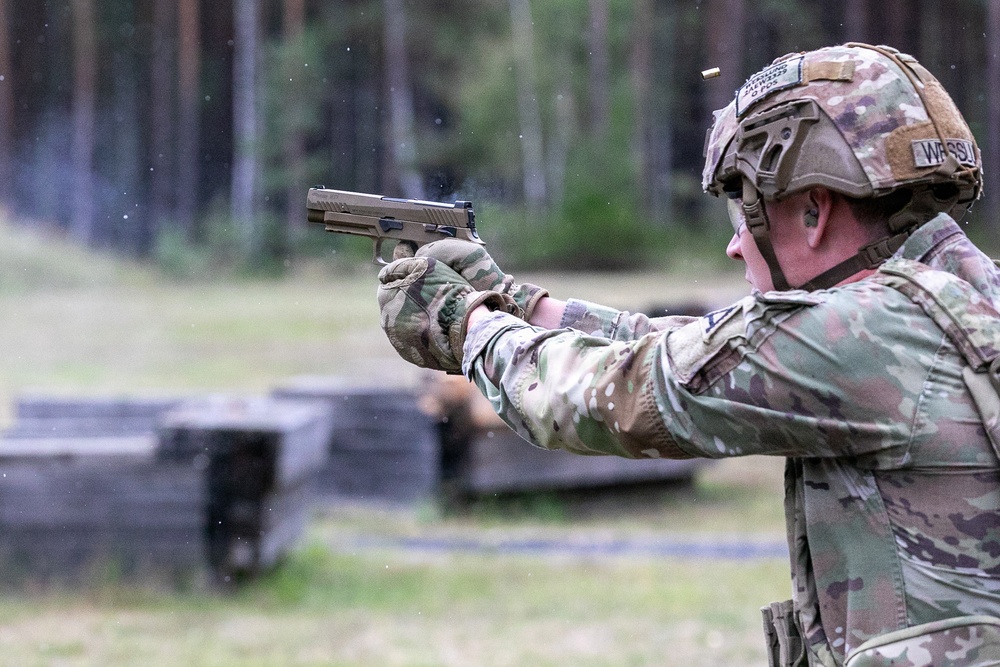 A military service member in full combat gear is firing a semi-automatic pistol at an outdoor shooting range. A spent brass casing is visibly ejecting from the firearm, indicating the weapon has just been discharged. The setting appears to be a structured training range with barriers visible in the background.