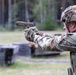 A military service member in full combat gear is firing a semi-automatic pistol at an outdoor shooting range. A spent brass casing is visibly ejecting from the firearm, indicating the weapon has just been discharged. The setting appears to be a structured training range with barriers visible in the background.