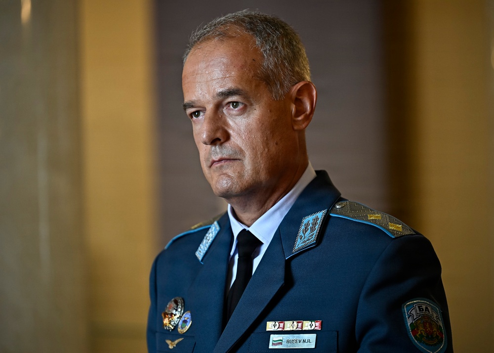 A military officer in a dark teal/blue dress uniform is photographed in a formal portrait-style setting. His name tag reads 'RUSEV N.A.' and he wears Bulgarian Armed Forces insignia, including a shoulder patch with the Bulgarian flag. He has a serious expression and is wearing multiple service ribbons and decorations.
