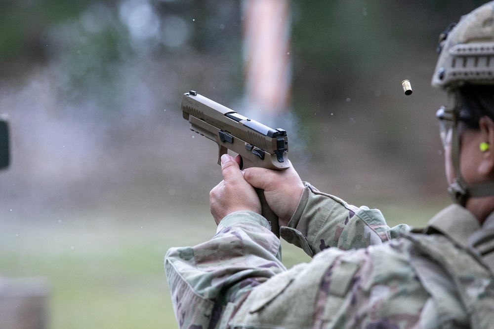 A military service member in full combat gear is firing a semi-automatic pistol at a shooting range. A spent brass casing is visibly ejecting from the firearm, captured mid-air to the right of the weapon. The shooter is wearing MultiCam/OCP camouflage uniform, a tactical helmet, and ear protection.