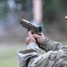 A military service member in full combat gear is firing a semi-automatic pistol at a shooting range. A spent brass casing is visibly ejecting from the firearm, captured mid-air to the right of the weapon. The shooter is wearing MultiCam/OCP camouflage uniform, a tactical helmet, and ear protection.