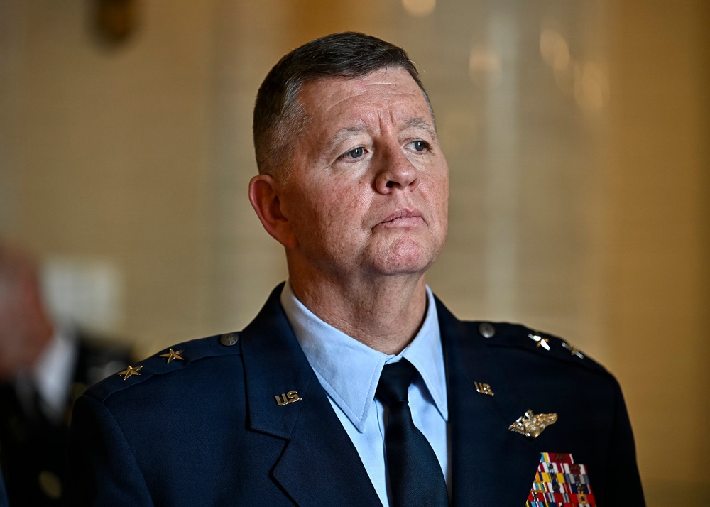 A senior U.S. military officer in dress uniform is photographed in what appears to be an official or governmental building. The individual is wearing a dark blue Air Force-style service dress uniform with multiple decorations and insignia visible. The expression is serious and attentive, suggesting a formal or official occasion.
