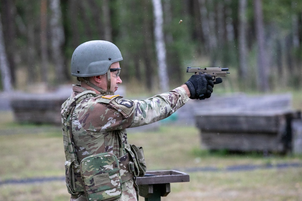 A U.S. Army soldier in full combat gear is firing a pistol at an outdoor shooting range. A spent brass casing is visibly ejecting from the firearm, captured mid-air above the weapon. The setting appears to be a military training range surrounded by a forested area.