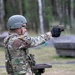 A U.S. Army soldier in full combat gear is firing a pistol at an outdoor shooting range. A spent brass casing is visibly ejecting from the firearm, captured mid-air above the weapon. The setting appears to be a military training range surrounded by a forested area.