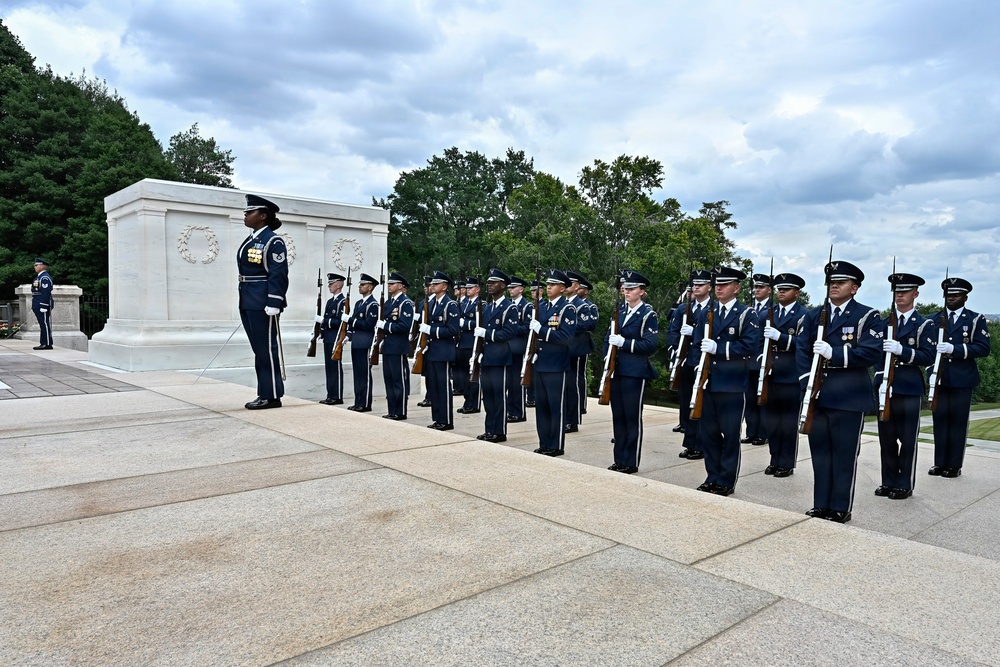A formal military honor guard ceremony is taking place at the Tomb of the Unknown Soldier at Arlington National Cemetery, Virginia. A large formation of U.S. Air Force personnel in dress blue uniforms stand at attention with rifles, flanking the white marble tomb. The ceremony appears to be a wreath-laying or changing of the guard event.