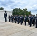 A formal military honor guard ceremony is taking place at the Tomb of the Unknown Soldier at Arlington National Cemetery, Virginia. A large formation of U.S. Air Force personnel in dress blue uniforms stand at attention with rifles, flanking the white marble tomb. The ceremony appears to be a wreath-laying or changing of the guard event.