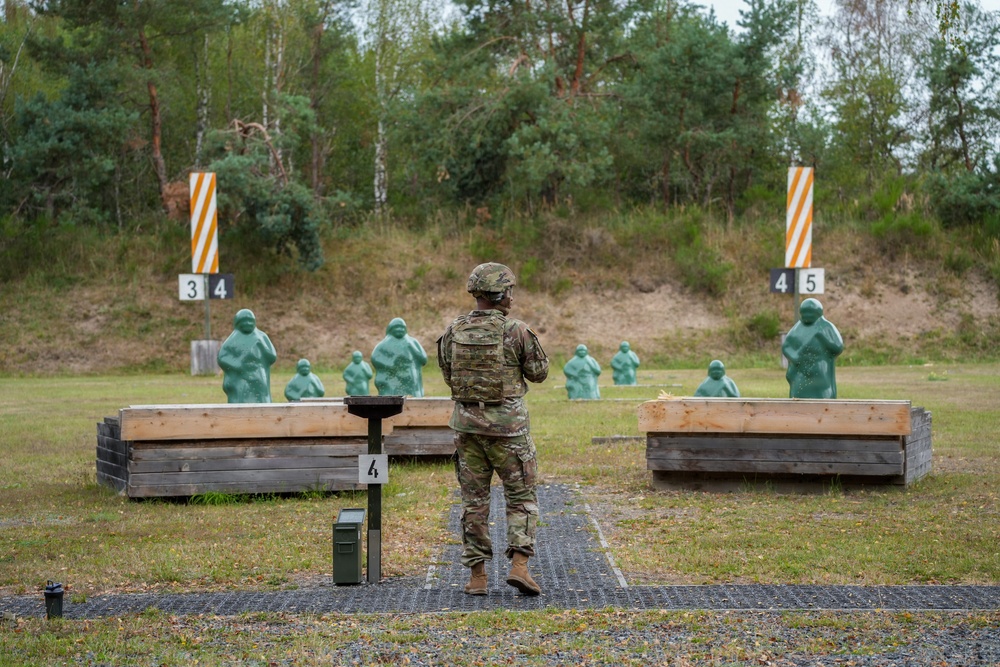 A soldier in full camouflage uniform and helmet stands at firing lane 4 of an outdoor military shooting range. Multiple green human-shaped silhouette targets are positioned downrange at varying distances. The range features wooden barricades, numbered lane markers, and is set against a backdrop of mixed forest.