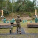 A soldier in full camouflage uniform and helmet stands at firing lane 4 of an outdoor military shooting range. Multiple green human-shaped silhouette targets are positioned downrange at varying distances. The range features wooden barricades, numbered lane markers, and is set against a backdrop of mixed forest.