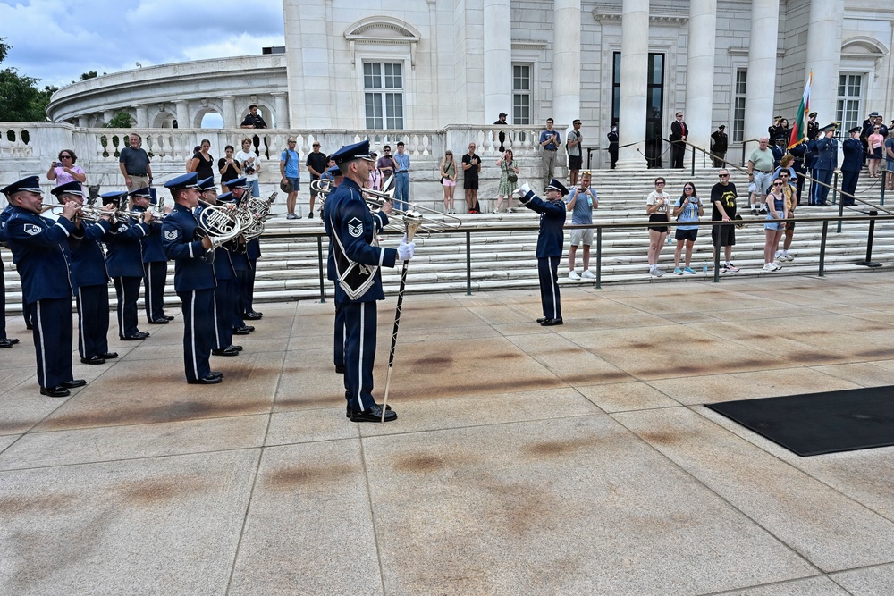 A military band, dressed in dark blue Air Force dress uniforms, performs outdoors on a stone plaza in front of a large white neoclassical building, likely the Memorial Amphitheater at Arlington National Cemetery. A drum major stands at center holding a ceremonial mace while musicians play brass instruments including French horns and trombones. Spectators and civilians line the steps and railings to observe the ceremony.