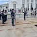 A military band, dressed in dark blue Air Force dress uniforms, performs outdoors on a stone plaza in front of a large white neoclassical building, likely the Memorial Amphitheater at Arlington National Cemetery. A drum major stands at center holding a ceremonial mace while musicians play brass instruments including French horns and trombones. Spectators and civilians line the steps and railings to observe the ceremony.